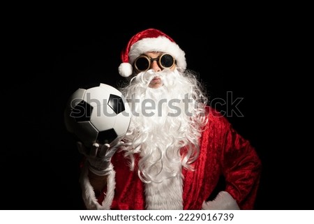 Santa Claus with sunglasses or a welder holding a World Cup soccer ball as a gift for all children, with a black background where his red suit and white beard stand out Royalty-Free Stock Photo #2229016739