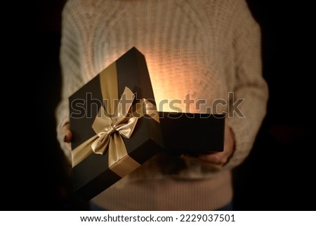 A girl in a sweater opens a black gift box with a golden bow. Open gift box with light from the inside.