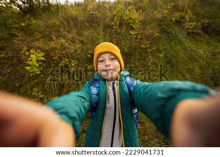 Cute boy making selfie,  wearing a green jacket, yellow hat, with backpack.