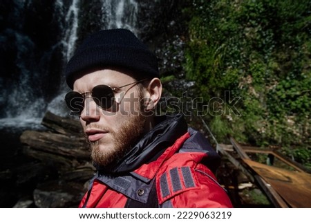 male traveler with a beard on a hike against the backdrop of a waterfall in the mountains