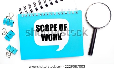 Office table with blue notepad, paper clips, magnifying glass and white card with text SCOPE OF WORK. Top view, flat lay. Stylish workplace
