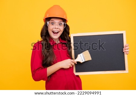 Painter in workshop. Renovation and repair. Teen girl in helmet with painting brush. Child in hard hat. Kid builder painter on construction site. Excited teenager girl.