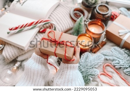 Hands in cozy sweater opening christmas gift with red bow on background of christmas tree with lights. Stylish female holding present with red ribbon in festive room close up. Merry Christmas!