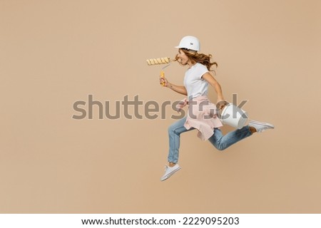 Full size young employee laborer handyman woman in white t-shirt helmet jump high hold paint roller bucket run isolated on plain beige background Instruments accessories renovation room Repair concept