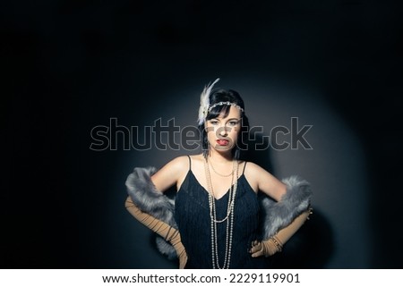 Studio portrait of a beautiful caucasian woman wearing flapper attire. She is standing in a patch of light. She is wearing a fringe trim dress, with elbow length gloves and a blue fur scarf.  Royalty-Free Stock Photo #2229119901