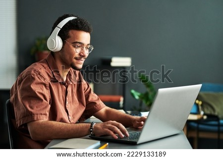 Young smiling man in headphones typing on laptop keyboard while sitting by workplace and taking part in online webinar or lesson Royalty-Free Stock Photo #2229133189