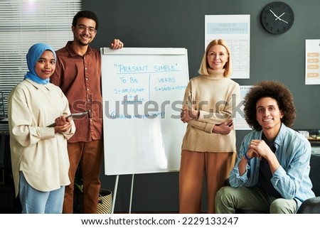 Several happy young intercultural students and mature blond teacher surrounding whiteboard with explanation of Present Simple tense Royalty-Free Stock Photo #2229133247