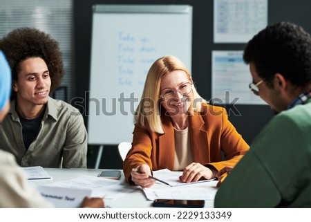 Happy mature blond teacher of linguistics explaining something to one of students while looking at him and pointing at document Royalty-Free Stock Photo #2229133349