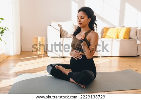 Pregnant african woman in lotus pose meditating on floor sitting on mat, doing pranayama breathing exercises for healthy pregnancy, preparing body for childbirth, putting hands of chest and belly Royalty-Free Stock Photo #2229133729
