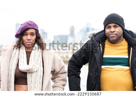 Portrait of two deaf friends looking at camera beside River Thames. It is Winter or Autumn, they are wearing coats and we can see skyscrapers behind them.