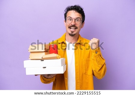 Young caucasian man holding fast food isolated on purple background celebrating a victory in winner position