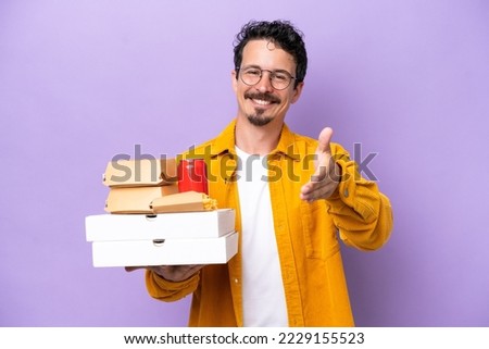 Young caucasian man holding fast food isolated on purple background shaking hands for closing a good deal
