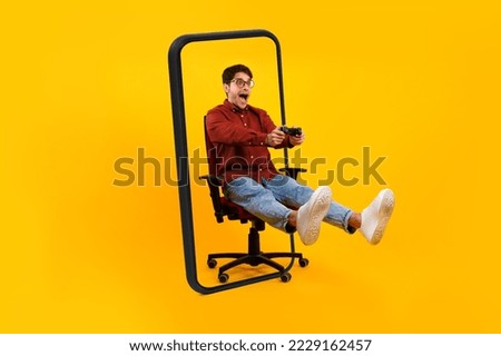 Happy surprised young european man gamer with open mouth in glasses sits on chair with joystick and plays in online game in huge phone isolated on yellow background. App and gadget, tech and emotions
