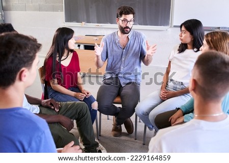 Diverse group of high school students sitting on chairs in a circle and interacting during a lesson, their Caucasian male teacher with them and talking. Education concept. High quality photo