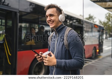 One man young adult male stand at public transport bus station taking bus with headphones and mobile smart phone in winter or autumn day with backpack student tourist city life copy space happy smile Royalty-Free Stock Photo #2229168201