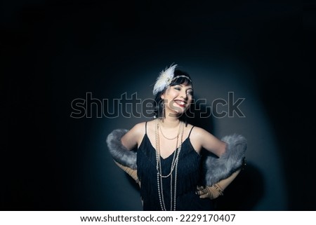 Studio portrait of a beautiful caucasian woman wearing flapper attire. She is standing in a patch of light. She is wearing a fringe trim dress, with elbow length gloves and a blue fur scarf.  Royalty-Free Stock Photo #2229170407