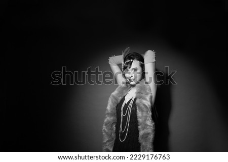 Black and white studio portrait of a beautiful caucasian woman wearing flapper attire. She is standing in a patch of light. She is wearing a fringe trim dress, with elbow length gloves and a headpiece Royalty-Free Stock Photo #2229176763