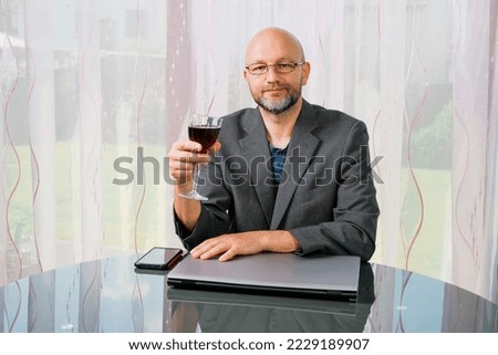 Bald businessman sitting at a table with notebook computer and glass of red wine. Working remotely and be your own boss concept. The model is in his 40s grey beard and suit. Office at home theme.