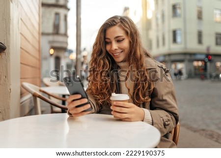 Young blonde woman in autumn coat sitting with smartphone in street cafe Royalty-Free Stock Photo #2229190375