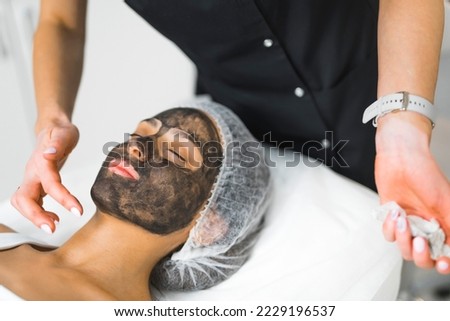 Relaxed Latin woman in her 30s lying down on SPA bed in protective hair cap when unrecognizable dermatologist in black uniform spreads charcoal mask on her face. High quality photo