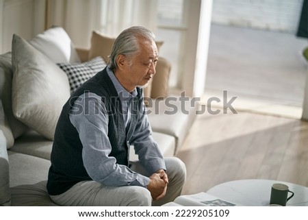 sad asian senior man sitting on couch in living room at home Royalty-Free Stock Photo #2229205109