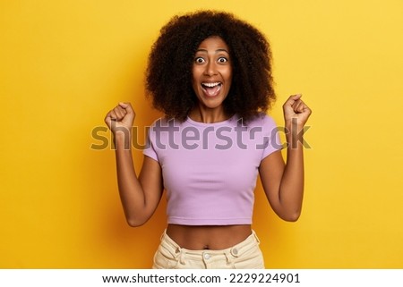 Positive excited brunette ethnic woman with wide opened mouth, clenches fists in winner gesture poses over yellow background. 