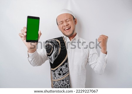 A happy young Asian Muslim man with a successful expression showing copy space on his phone isolated by white background