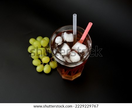 A tall glass of fruit juice with ice and straws on a black background, next to a bunch of white grapes. Top view, flat lay.