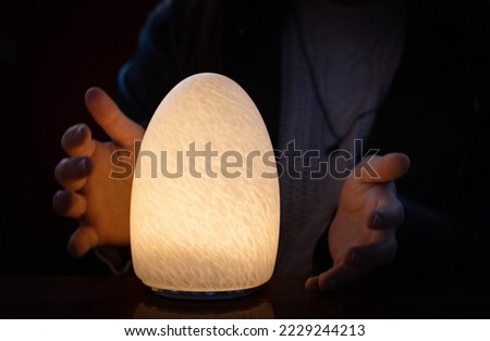 A night lamp in the shape of an egg with an abstract and textured surface is lit with a yellow warm light and the hands of a young girl are trying to grasp it, side view close-up.