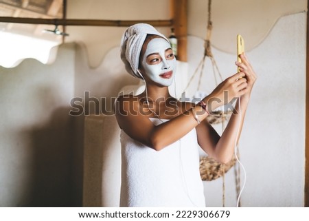 Young Asian female shooting beauty vlog while doing mask procedures for nourishing treatment in home bathing using front camera on digital cellular, attractive woman in earphones making selfie photos