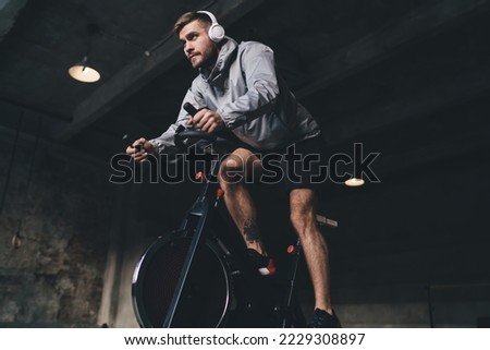 Low angle of young male in headphones looking away while sitting on spinning stationary cycle and listening to music in wireless headphones while doing cardio workout in gym Royalty-Free Stock Photo #2229308897