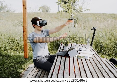 Young man sitting on wooden bench with virtual reality glasses on. On the table are work documents, notebook, plants and in the background is an e-scooter.