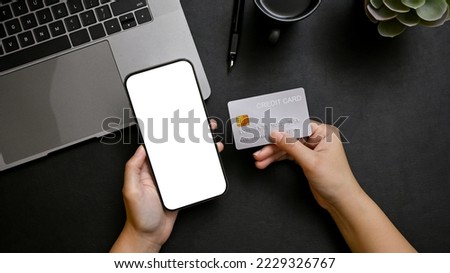 Top view, A female hands holding a smartphone white screen mockup for display your graphic ads and credit card over modern black office desk. online payment, mobile banking, online shopping
