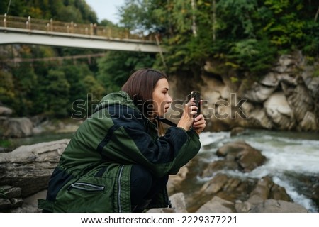 Female tourist sits on a rock near a mountain river and takes a photo on a smartphone camera with a serious face