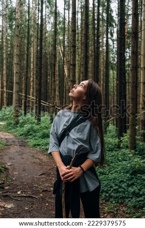 A woman tourist with a stick in her hands stands in a pine forest and looks up at the beautiful trees. Vertical photo