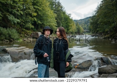 Photo of two stylish young tourists standing on a rocky bank of a mountain river in the background of a beautiful landscape and talking.