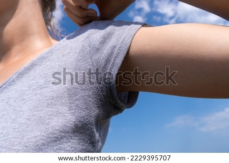 Close-up of a woman with her hand raised up in a gray T-shirt on which sweat is visible on a sunny summer day against a blue sky