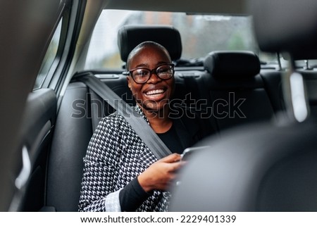 A cheerful African American woman is in the car on the backseat using her smartphone and looking at the camera. Royalty-Free Stock Photo #2229401339
