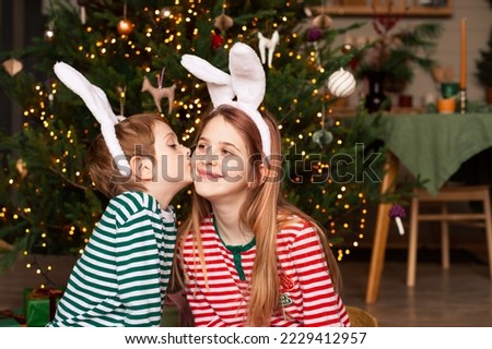 Little boy kissing his sister wearing striped pajamas and rabbit ears near christmas tree. Happy children with festive freckles tattoos waiting for the New Year of the Rabbit. Selective focus.