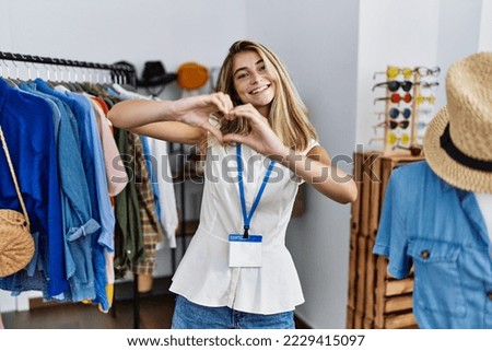 Young blonde woman working as manager at retail boutique smiling in love doing heart symbol shape with hands. romantic concept. 