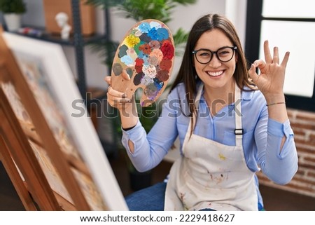 Young brunette woman holding painter palette at art studio doing ok sign with fingers, smiling friendly gesturing excellent symbol 