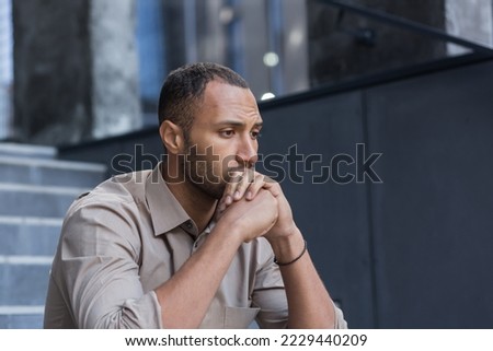 Portrait of a serious and sad hispanic.african american man. He sits sullenly outside on the stairs, holds his head in his hands, looks to the side. Royalty-Free Stock Photo #2229440209