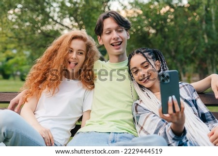Portrait of a happy three teenagers sitting on the bench and making photo on smartphone. Relaxing on the bench in university campus. Sunny summer day.