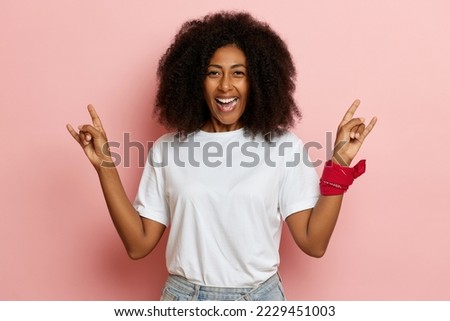 Young positive curly haired dark skinned woman smiles and shows horn gesture, she stands isolated over pink wall