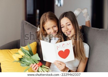 Child daughter congratulates mother and gives present card, gift and bouquet of flowers at home. Mom and girl smiling and hugging on couch. Happy mothers day celebration