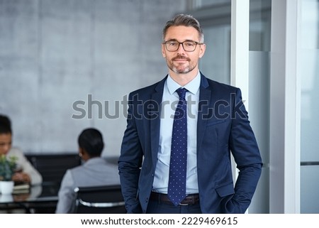 Portrait of mid adult business man standing in modern office. Successful mature entrepreneur in formal clothing looking at camera with satisfaction. Confident man in suit with eyeglasses and beard. Royalty-Free Stock Photo #2229469615