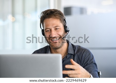 Technical support operator working with headset in office. Smiling handsome man working as call centre operator, speaking to customer. Happy businessman working remotely while doing video conference. Royalty-Free Stock Photo #2229469629