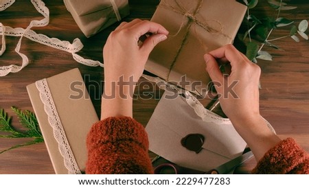 Woman making a ribbon on a kraft box with a sealed wax letter between the arms.Wood background. Aerial shot
