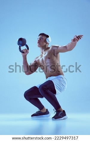 Portrait of young muscular man in headphones training, lifting weight and doing squats isolated over blue background in neon light. Concept of sport, fitness, healthy and active lifestyle, motivation