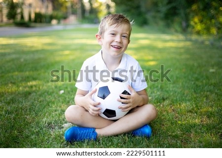 a little blonde boy is sitting on the grass with a soccer ball in the summer, looking at the camera